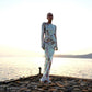 A woman standing on a platform at sunset wearing a sequined, silver, maxi dress with a one-shoulder design and crew neckline.