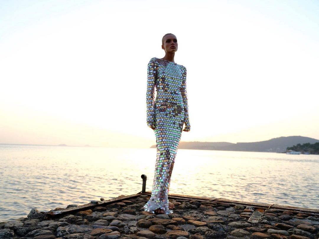 A woman standing on a platform at sunset wearing a sequined, silver, maxi dress with a one-shoulder design and crew neckline.